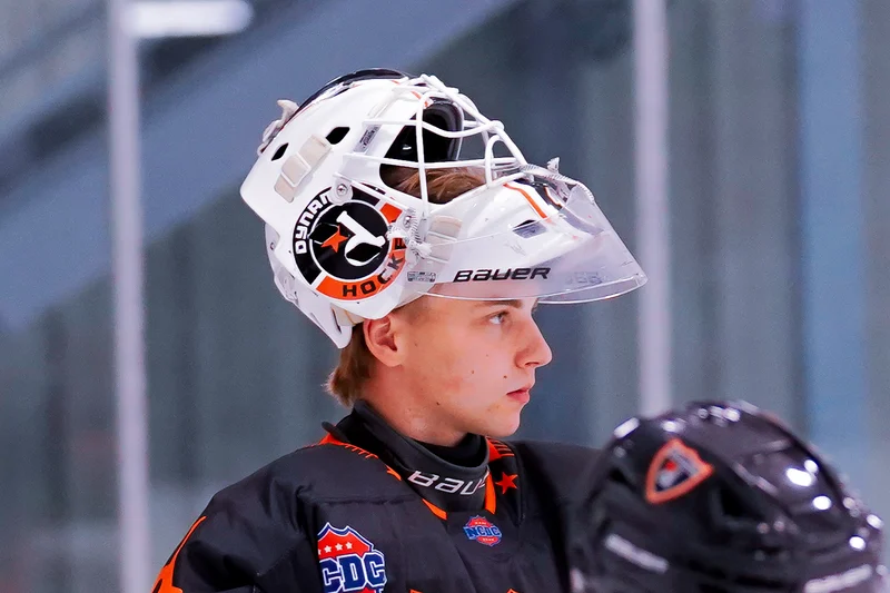 A hockey goalie stands at ease, in full gear, with his helmet perched up to offer a full view of the rink.